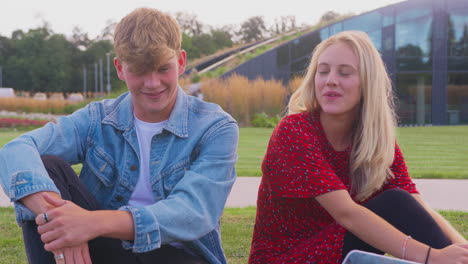 male and female university or college students sit on grass outdoors on campus talking and laughing