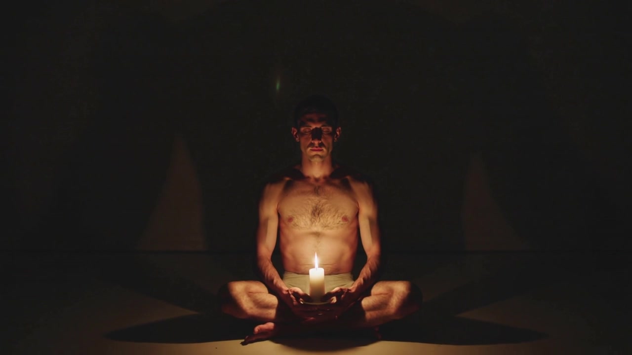 Holding lit candle in bowl as flame flickering, shirtless man in shorts sitting on mat meditating
