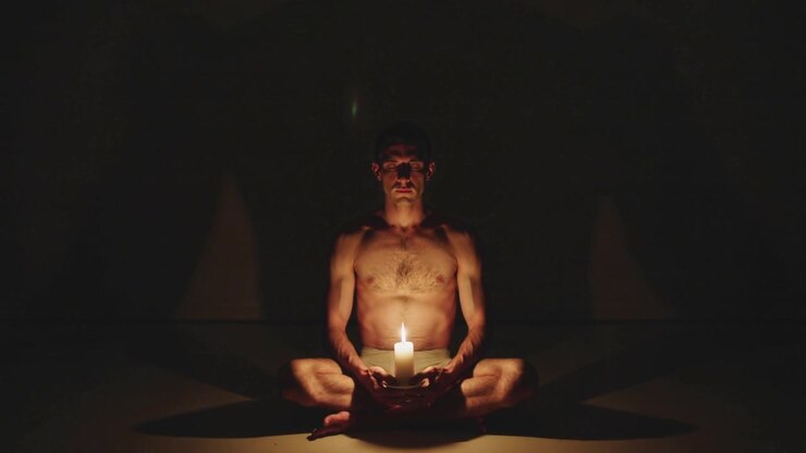 Holding lit candle in bowl as flame flickering, shirtless man in shorts sitting on mat meditating
