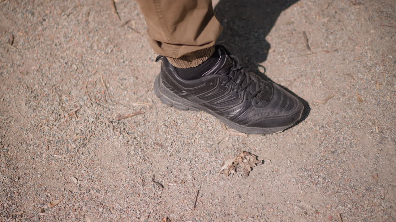 Closeup Shoe Stomps Empty Plastic Bottle On Dusty Ground, Forceful Downward Motion Flattens Debris, Khaki Trousers And Shadow Frame Action, Sunlight Reveals Crushed Contours, Scene Conveys Cleanup
