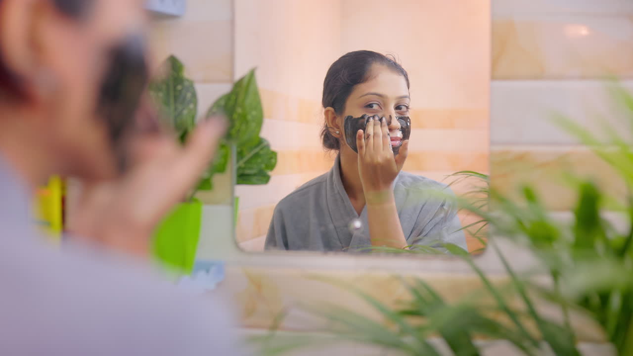 Young female applying charcoal facial mask in front of a bathroom mirror during her skincare routine at home