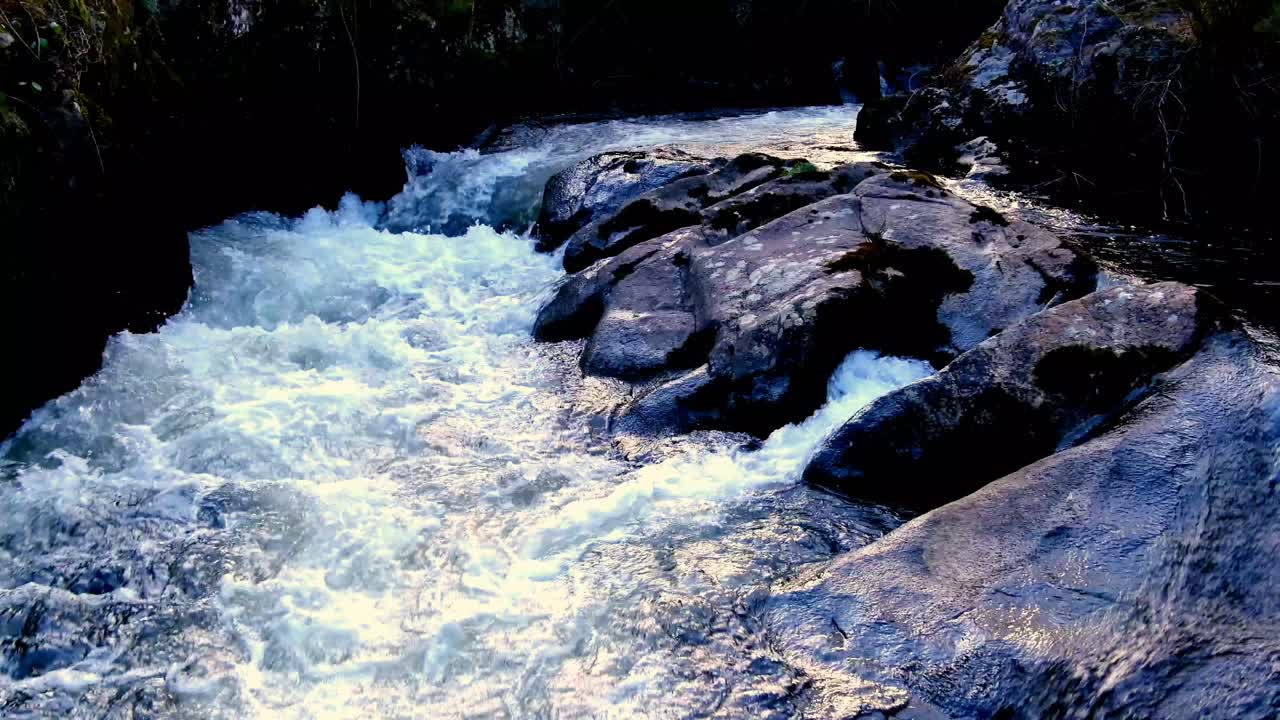 Acqua di fiume che scende a cascata su rocce scure. Dolly in avanti.