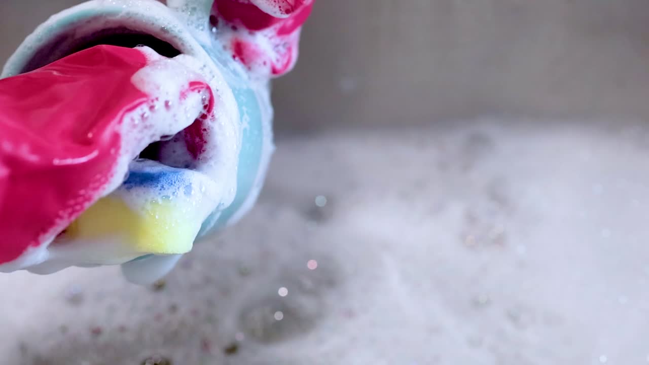 A person wearing pink rubber gloves washes a blue ceramic cup in soapy sink water