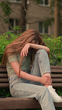 dama en vaqueros casuales y zapatillas blancas se sienta en un banco de madera en el parque, descansando la cabeza sobre el brazo en una postura reflexiva y sombría con el cabello en cascada por el hombro, rodeada de exuberante vegetación y árboles