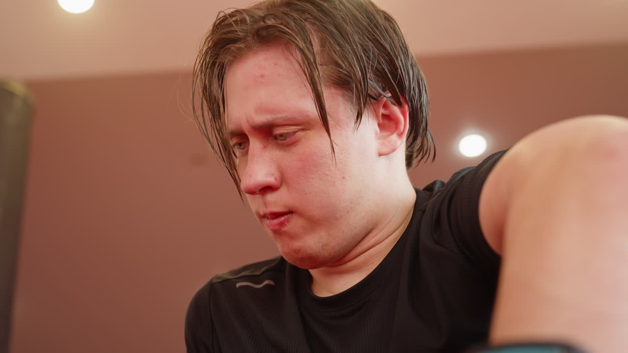 Male fighter breathing heavily after sparring in gym, face flushed with effort, sweat visible, wearing black sportswear, showing exhaustion, struggle, determination under bright lights during intense training