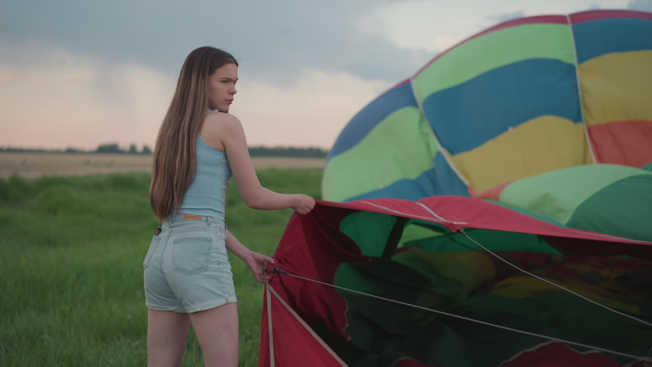 young woman holding vibrant hot air balloon envelope panels by sturdy cables while preparing for sunrise flight in grassy field under soft pastel sky reflecting morning light on fabric textures