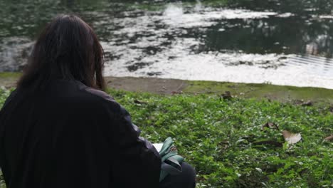 View-Behind-Young-Girl-Sketching-At-The-Park-While-Sitting-On-Grass