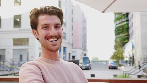 Millennial-white-male-hipster-sitting-in-a-city-street-laughing-to-camera,-close-up