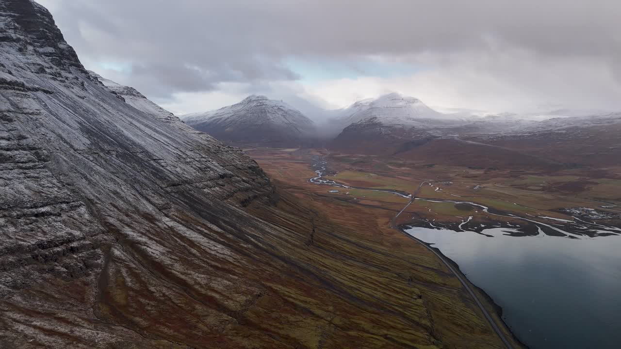 Ilmakuva Islannin Reyðarfjörðurista pilvisenä päivänä. Lumihuippuisia vuoria ja mereen virtaava joki.