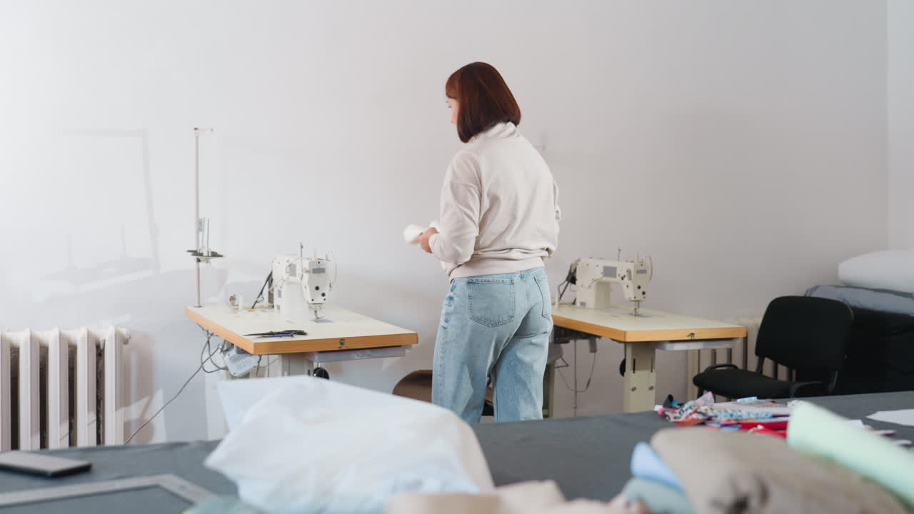 Seamstress wearing jeans and plain long sleeve top taking white thread from large table walking to industrial sewing machine placing thread on machine ready to sew in spacious tailoring workshop