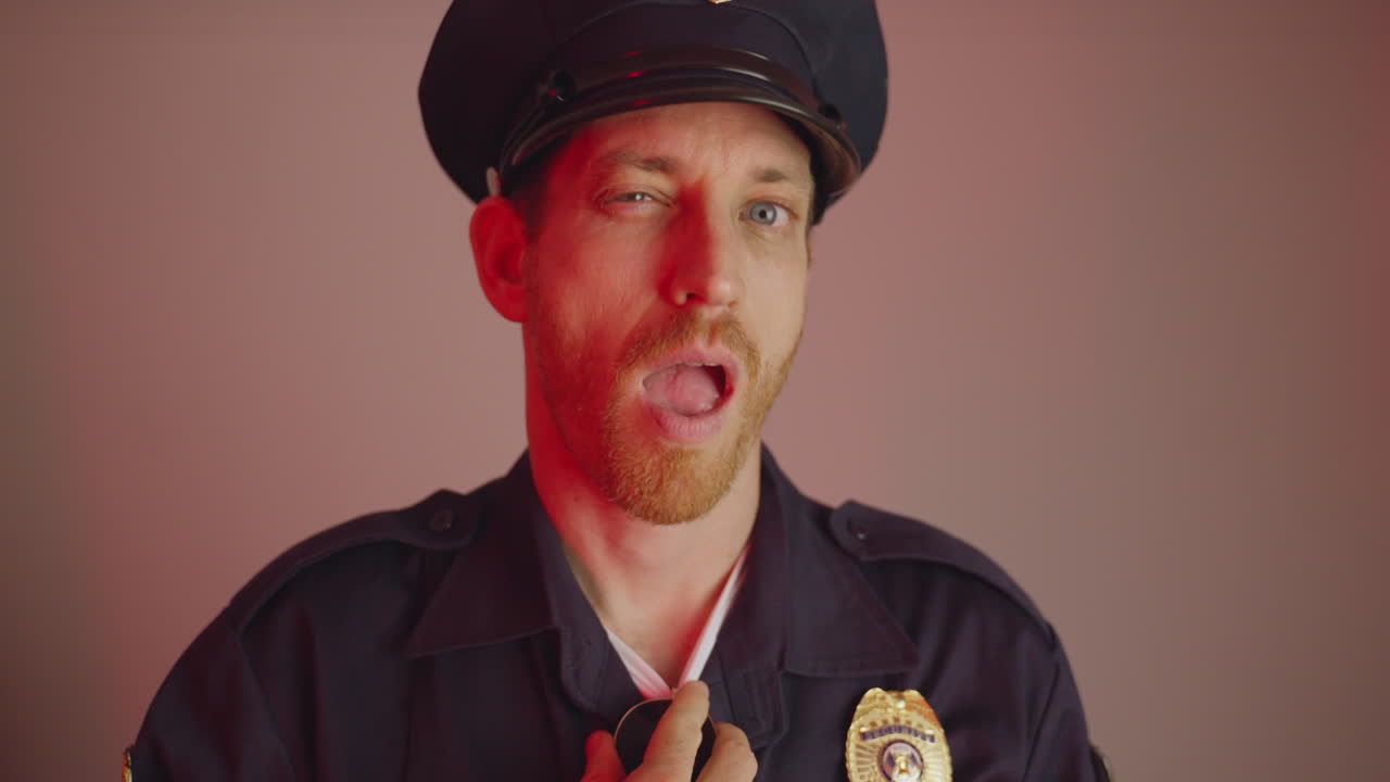 Serious looking cop or policeman takes off sunglasses and winks to camera. Portrait of police officer with blue uniform and flashing blue and red lights smiles and winks in humor joke or in on idea