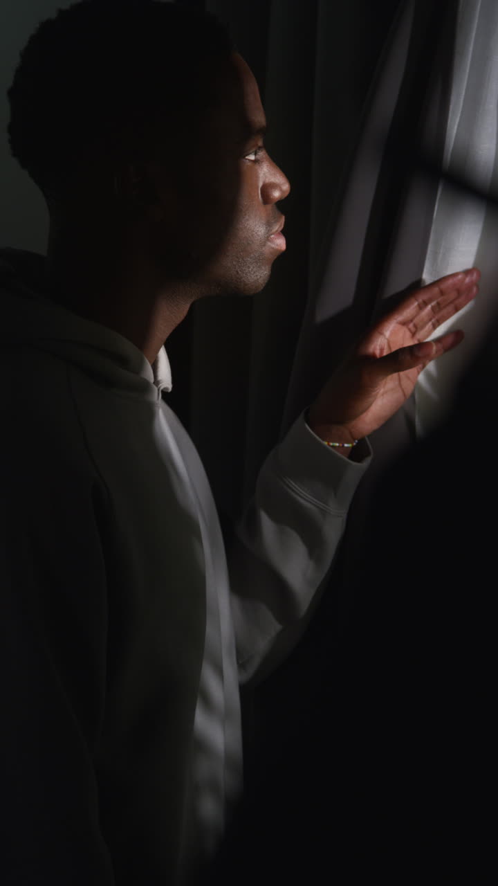 video vertical de un joven infeliz y deprimido en casa mirando por la ventana sufriendo de agorafobia o paranoia 1