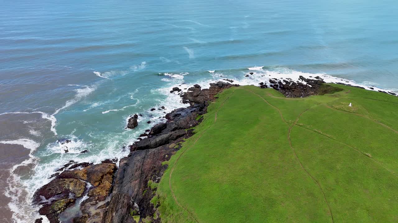 Smooth drone flyover reveals lush green headland, rugged cliffs, and ocean waves under daylight