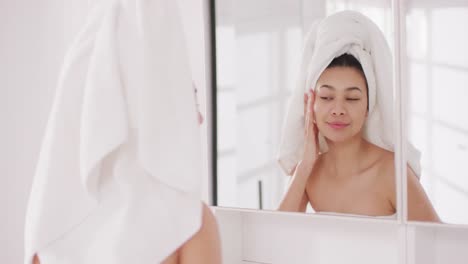 video of portrait of smiling biracial woman with towel on hair looking in mirror in bathroom