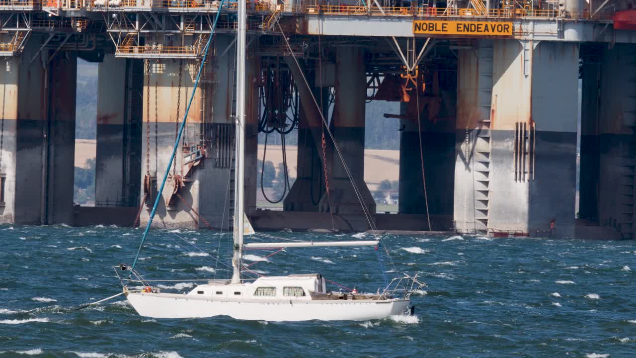 White sailboat moves past large offshore oil platform in daylight, choppy sea, telephoto perspective