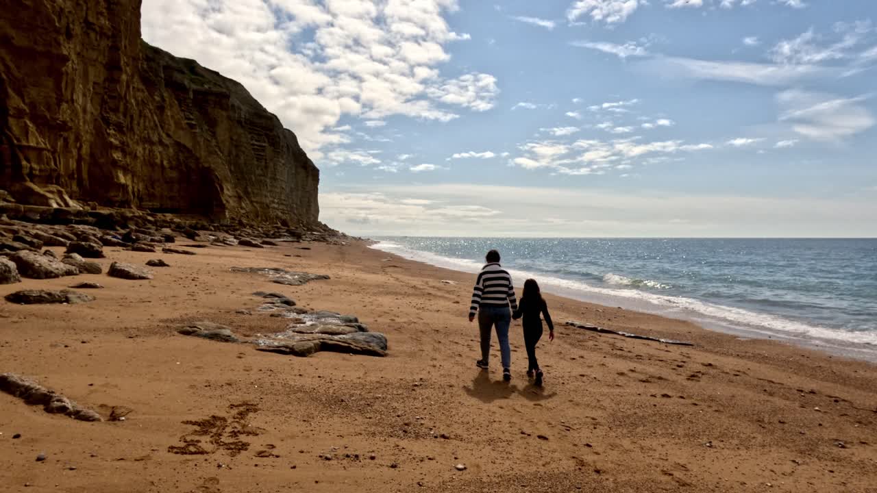 A mum and daughter walking along a Jurassic Coast beach in Dorset. Captured in slow motion the girls come into shot from the right and walk away along the beach with a tall cliff face on the left.