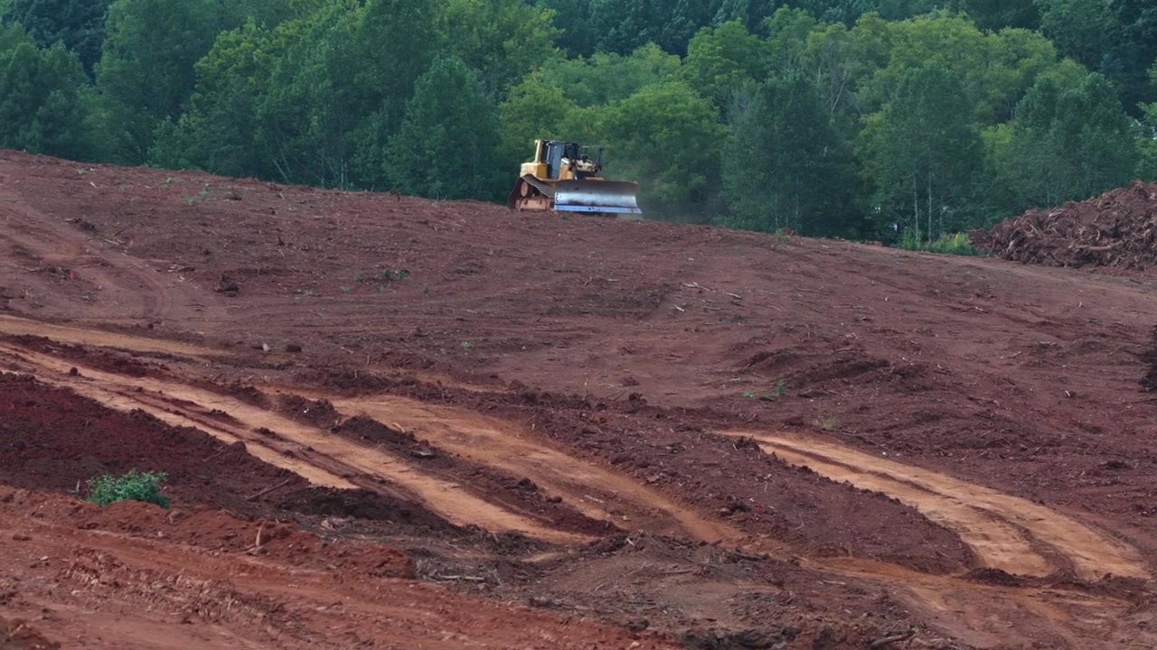 Bulldozer jævner ryddet bjergskråning nær Old Graves Mill Road, dækspor skærer sig gennem rød lerjord, tæt skov omkranser højderyg over nyjævnet byggegrund. Bredt luftfoto med drone.