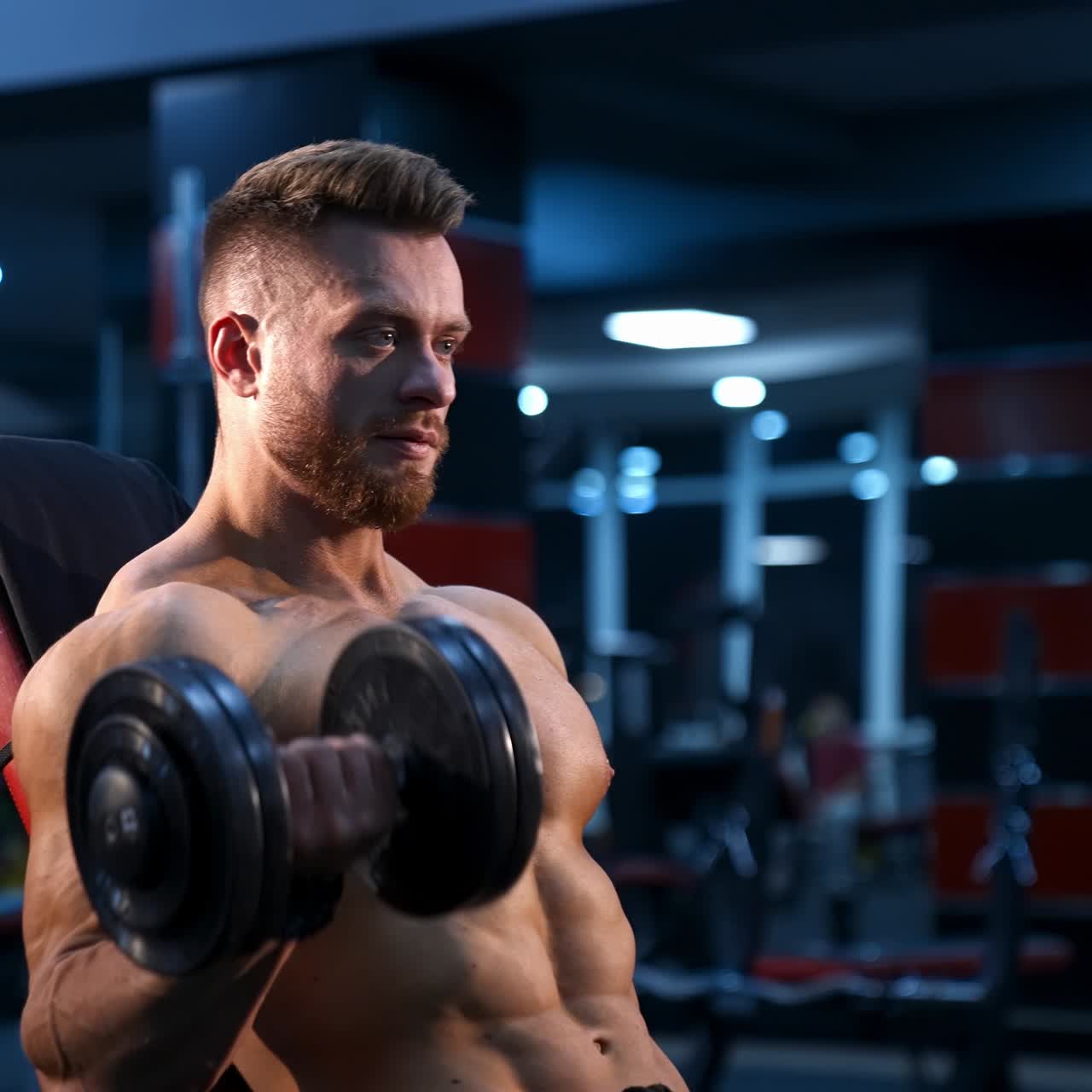 Shirtless bodybuilder in gym. Young guy with muscular body is lifting dumbbells. Sportsman practicing at sports center while sitting. Hard strength workout. Profile view.