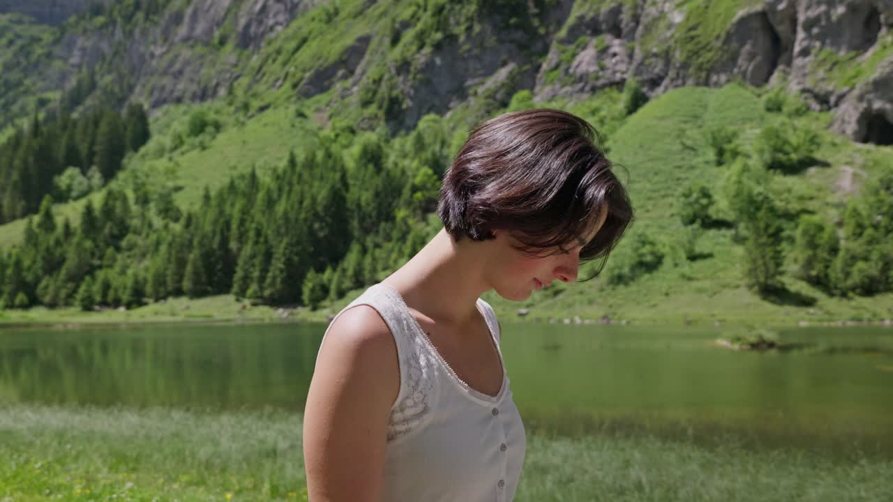 A smiling woman walks near a peaceful lake with lush mountains and trees in the background