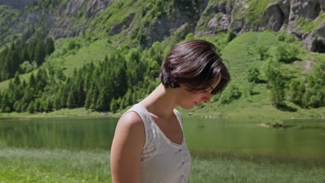 a smiling woman walks near a peaceful lake with lush mountains and trees in the background