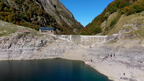 Lac-d'Oô-artificial-lake-in-the-French-Pyrenees-with-visitors-scaling-the-dam-rock-wall-ridge,-Aerial-pan-left-shot