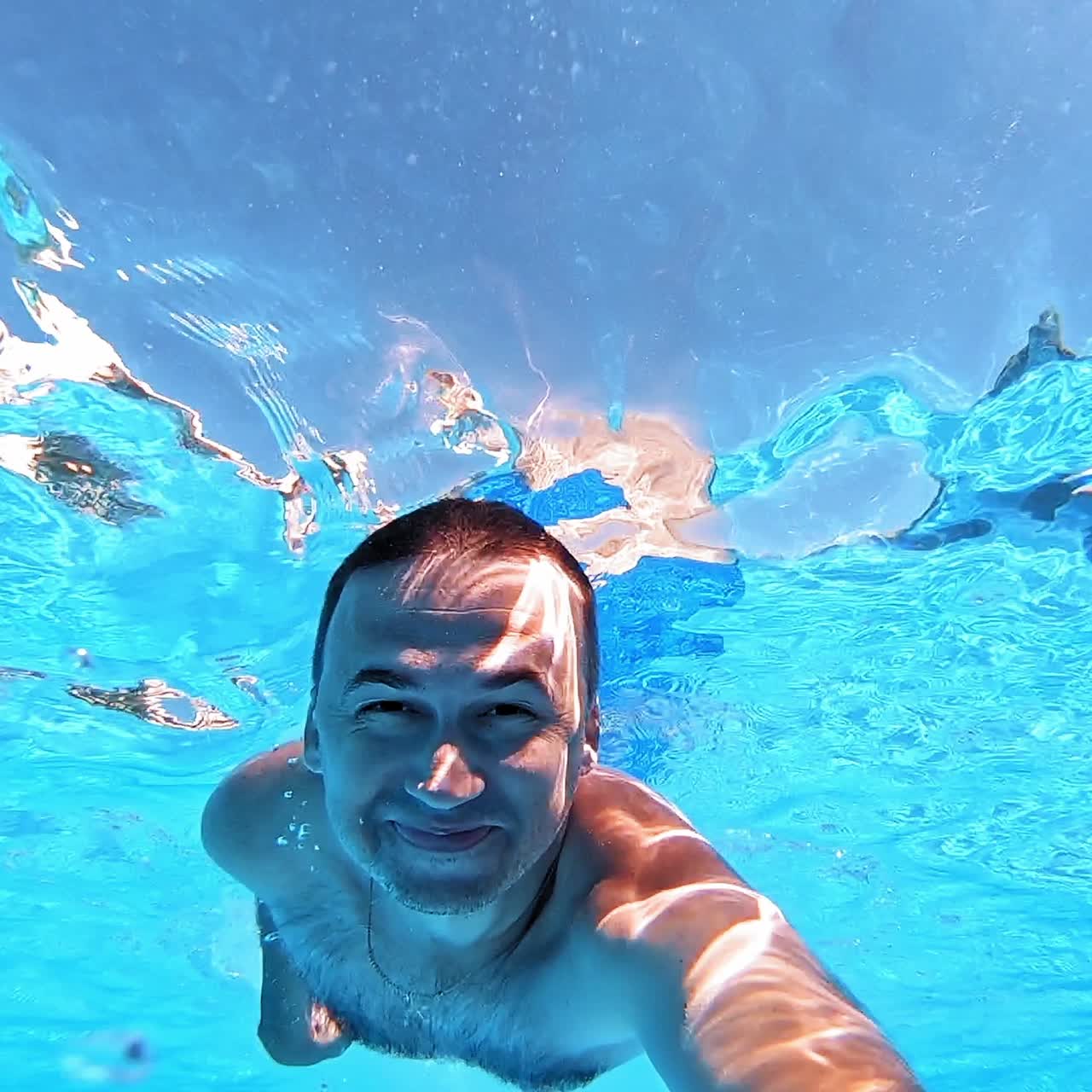 Man smiling on camera underwater in the pool. Happy man dives under the blue clear water and swimming inside the swimming pool in summer.
