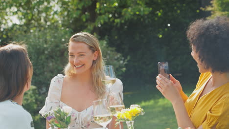 woman taking photo of friends sitting outdoors in summer garden drinking wine and making a toast