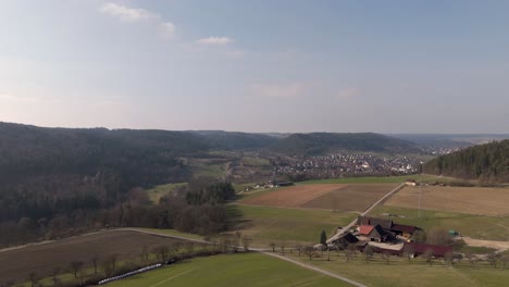 Aerial-dolly-forward-over-rural-landscape,-farmland-fields-with-village-in-background-in-valley-surrounded-by-hills