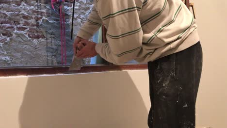 young man removing excess of plaster with scraper around wooden window frame with old brick wall in background in 4k