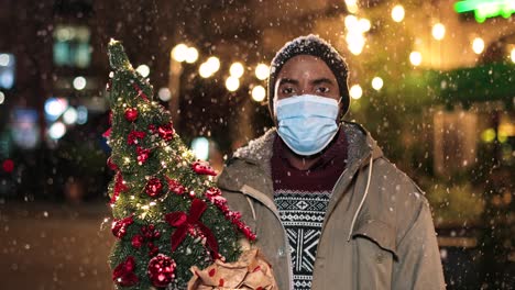 retrato de un joven afroamericano con máscara facial sosteniendo un árbol de navidad y sonriendo a la cámara en la calle mientras nieva en navidad