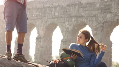 happy young couple backpackers tourists on a log trunk playing guitar singing reading book in front of ancient roman aqueduct ruins in romantic parco degli acquedotti park in rome at sunrise slow motion