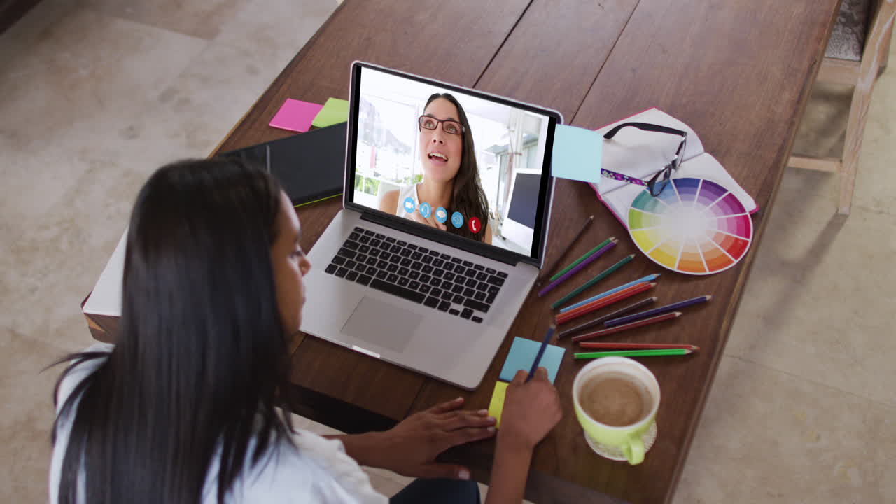 Mujer caucásica usando una laptop en videollamada con una colega y tomando notas