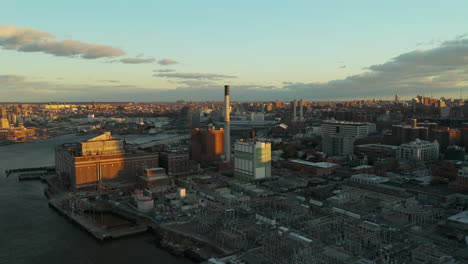 Aerial-view-of-industrial-site-on-riverbank.-Electricity-substation,-storage-facility-and-power-station-with-chimney.-City-lit-by-setting-sun.-Brooklyn,-New-York-City,-USA