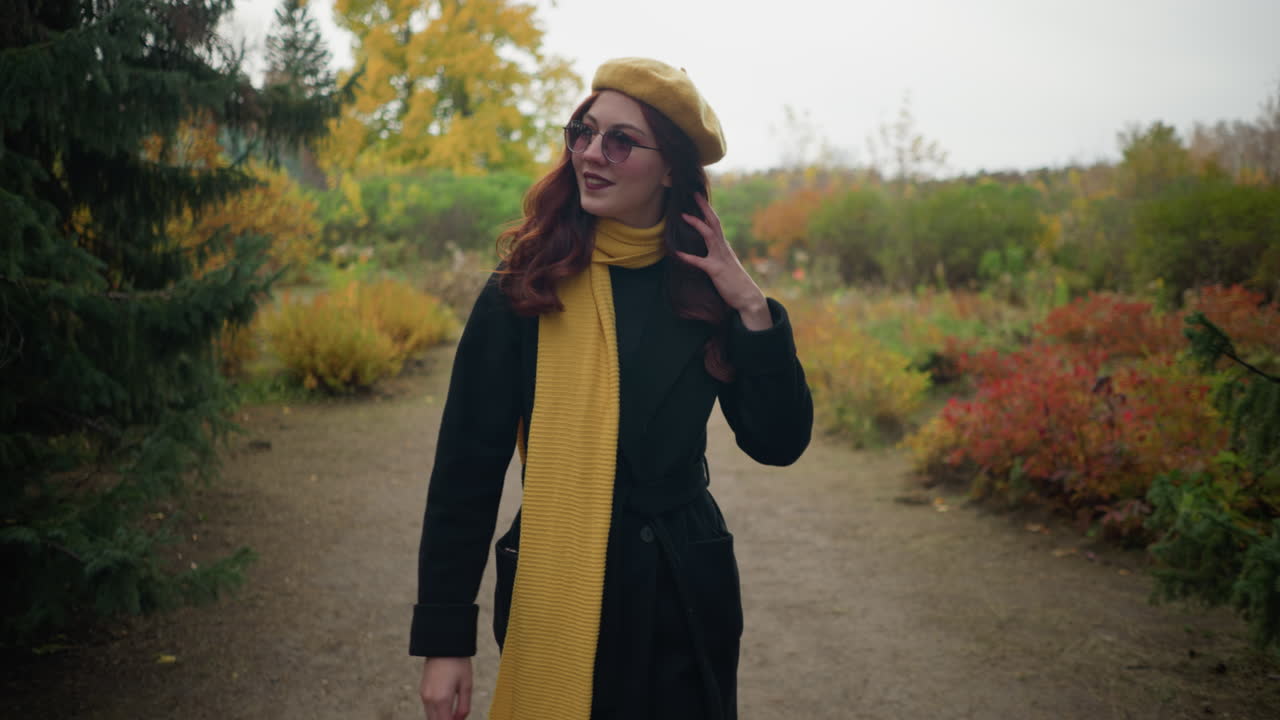 Studentessa con un basco giallo e occhiali da sole cammina attraverso un bellissimo giardino, toccandosi i capelli e sistemandosi gli occhiali, circondata da un vivace fogliame autunnale.