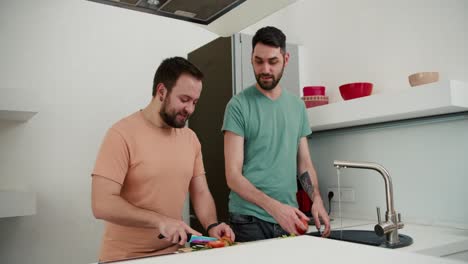 Una-Pareja-Gay-Ayuda-A-Preparar-La-Comida,-Uno-Lava-Las-Verduras-Y-El-Otro-Corta-Las-Verduras-En-La-Cocina.