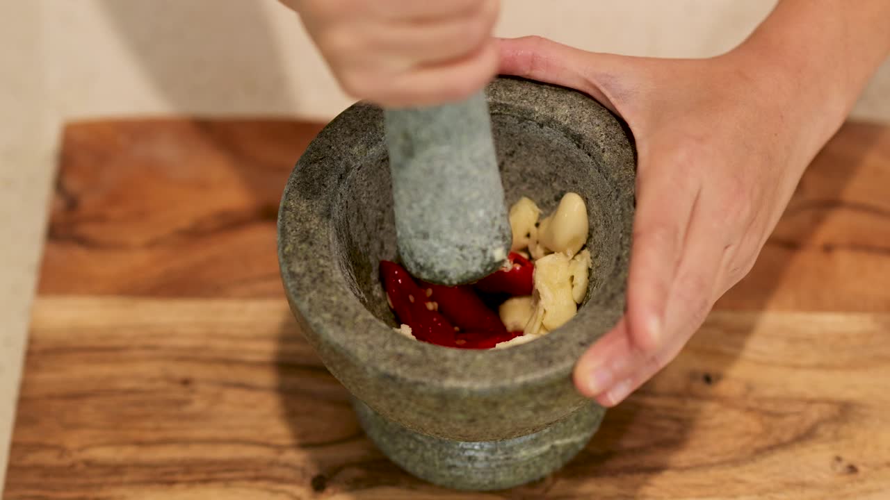 Hands using a stone mortar and pestle to crush garlic and chili on a wooden surface