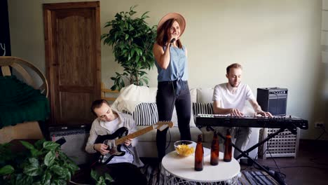 pretty blonde vocalist from musical band is singing and dancing while her friends guitarist and keyboarder are playing guitar and keyboard and smiling.