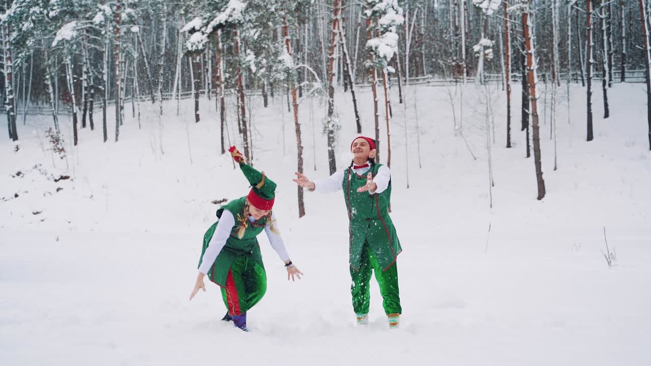 Funny elves in green suits with hats are throwing up snow and looking at it on the background of a winter park. Slow motion