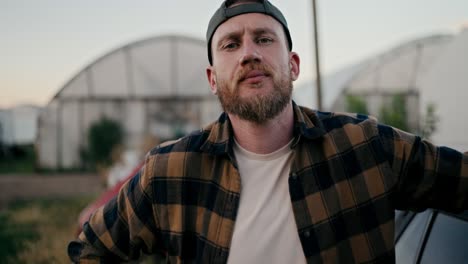 Portrait-of-a-confident-farmer-guy-with-a-beard-in-a-checkered-shirt-posing-near-the-car-among-the-greenhouses-on-the-farm