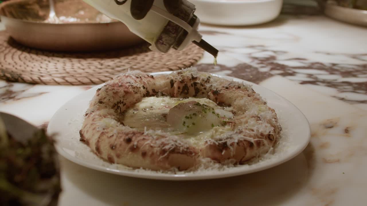 A detailed close-up shot of a chef's hand drizzling oil over a freshly baked Khachapuri, a traditional Eastern European cheese bread topped with a sunny-side-up egg
