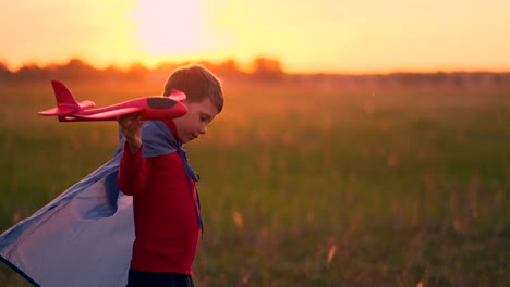 piloto de niño corre en un impermeable rojo sosteniendo un avión riendo al atardecer en el campo de verano imaginando que es un piloto de avión jugando con un modelo de avión