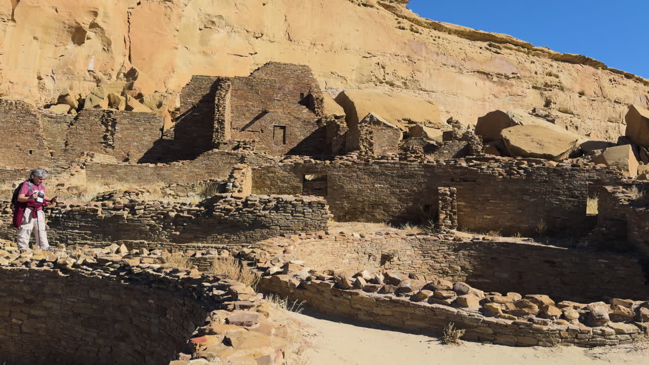 Some tourists walking among large stone ruins of a desert cliffside archaeological site in Chaco Canyon, New Mexico.