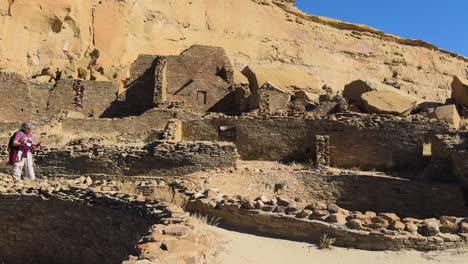 some tourists walking among large stone ruins of a desert cliffside archaeological site in chaco canyon, new mexico.