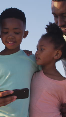 smiling african american family using smartphone and embracing on sunny beach