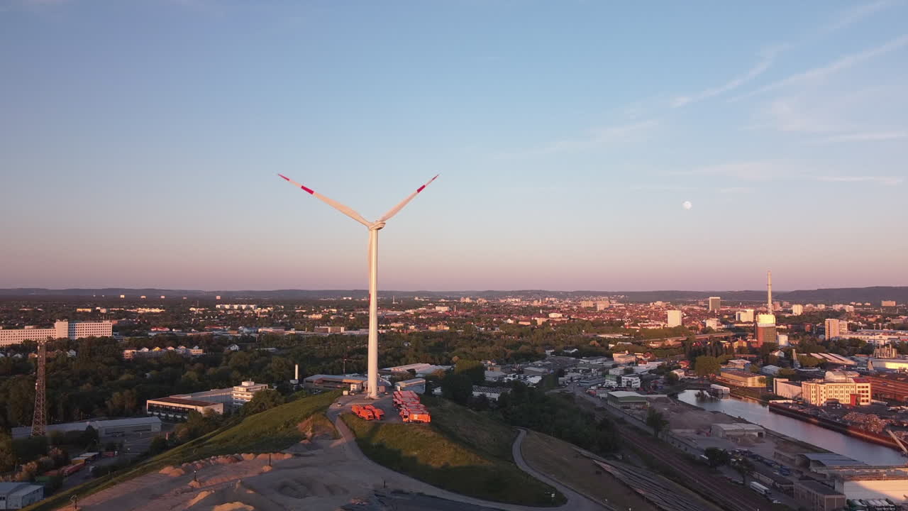 Drone flight toward a wind turbine in Germany sitting on a garbage dump with scenic city view in the background while sun is setting low