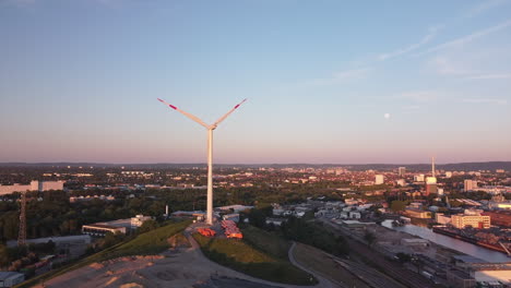 drone flight toward a wind turbine in germany sitting on a garbage dump with scenic city view in the background while sun is setting low