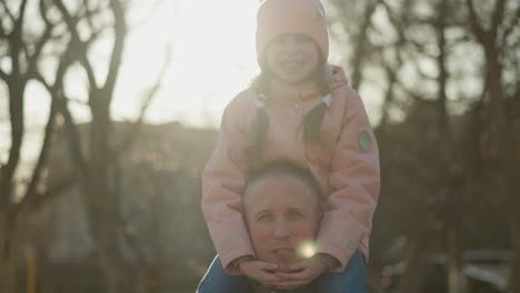 front view of a little girl in a pink cap and jacket sitting joyfully on her dad's neck as they walk through a sunlit park. the girl is smiling with her eyes closed