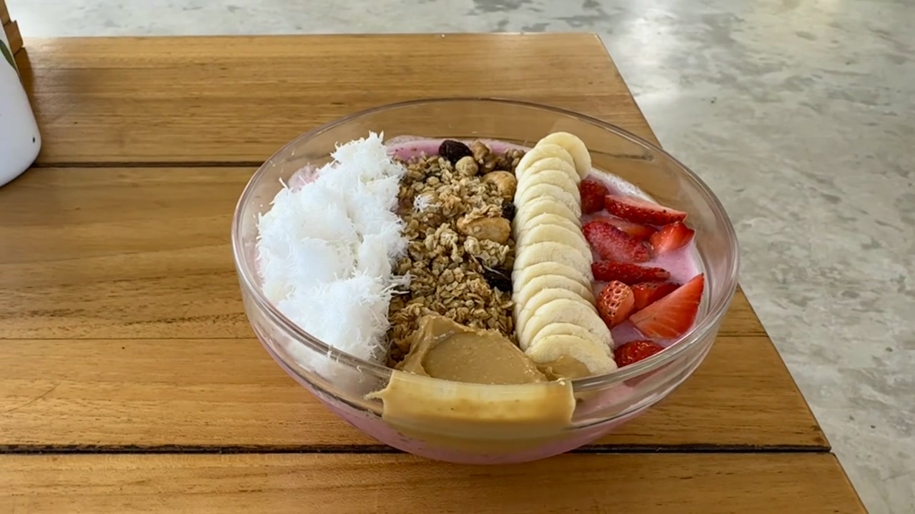 Lähikuvaa eloisasta lohikäärmehedelmä-smoothiekulhosta, jonka päällä on banaania, mansikkaa, kookoshiutaleita, kotitekoista taateligranolaa ja maapähkinävoita terveelliseksi aamiaiseksi.