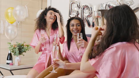 group of multiethnic female friends dressed in pink silk nightdresses talking and laughing while holding champagne glasses