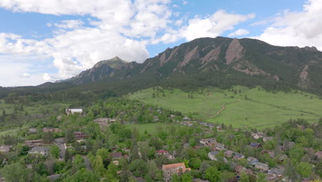 Wide-pan-right-aerial-view-of-warm-sun-hitting-Boulder-Colorado-Flatiron-mountains-above-Chautauqua-Park-with-full-green-pine-trees-and-blue-skies-with-clouds-on-a-beautiful-summer-day-for-hiking